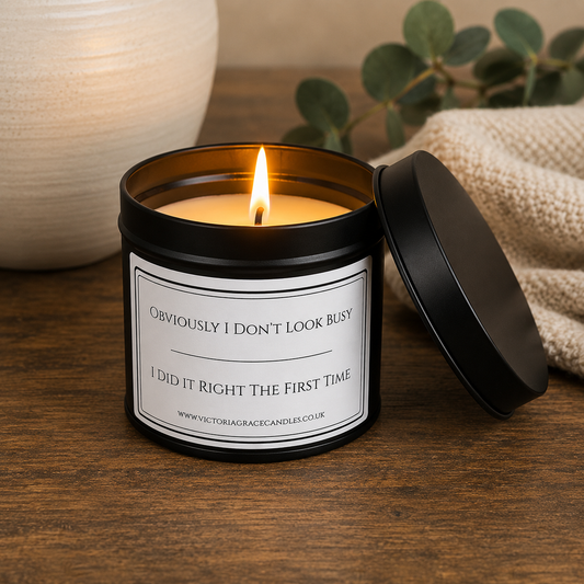 A lit candle in a black metal tin with a white label that reads 'Obviously I Don't Look Busy / I Did It Right The First Time.' The tin sits on a wooden surface with its lid leaning beside it, and a neutral home-style setting with a textured vase, cloth, and greenery in the background.