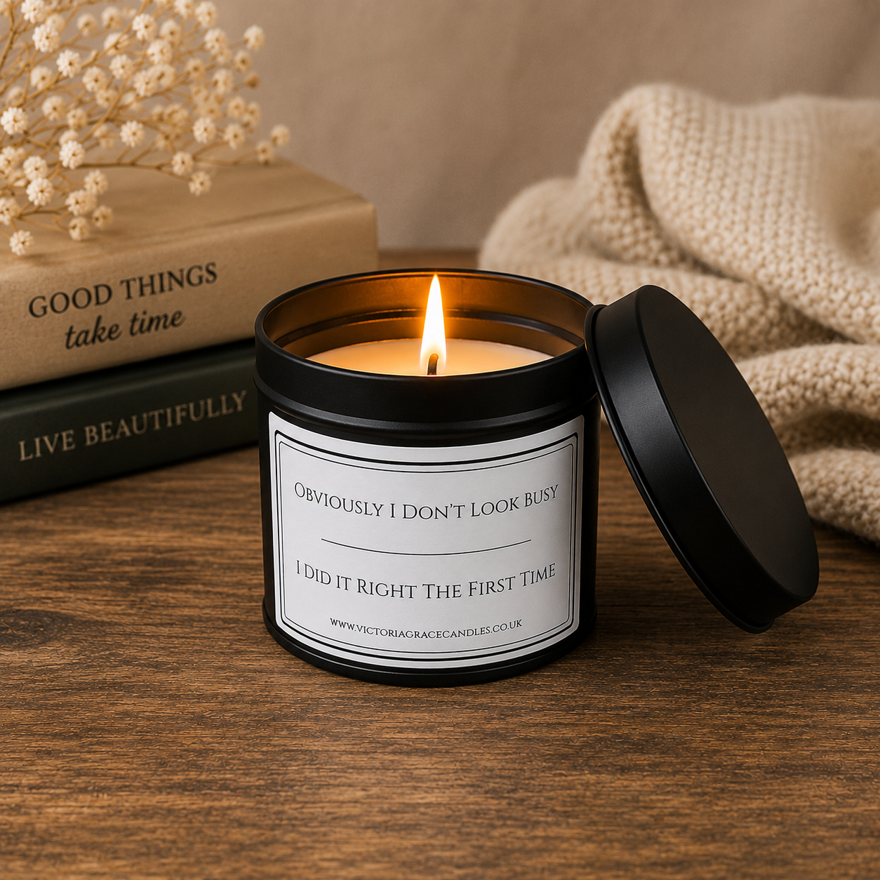 A lit candle in a black glass jar with its lid leaning beside it on a wooden surface. The candle has a white label with black text that reads, 'OBVIOUSLY I DON'T LOOK BUSY / I DID IT RIGHT THE FIRST TIME.