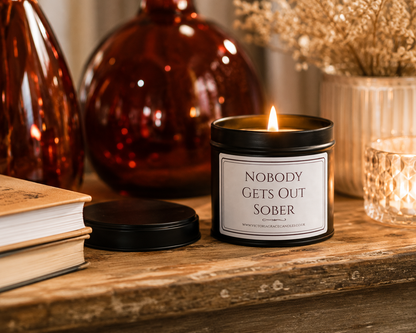 A lit black metal candle tin with a white label reading 'NOBODY GETS OUT SOBER' sits on a rustic wooden surface, with the lid placed beside it. The scene includes amber glass bottles, a book, and dried flowers in the background.