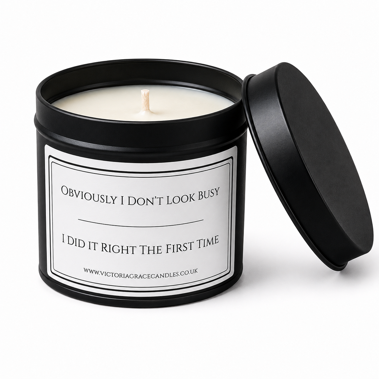 A black metal candle tin with its lid leaning against it, showing a white wax candle and a white label that reads, 'Obviously I Don't Look Busy / I DID IT RIGHT THE FIRST TIME' 